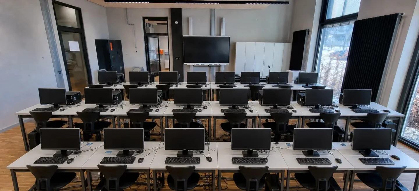 Klassischer Computerraum vor der Umrüstung: Tischreihen mit festen Monitoren, Tastaturen, Tower-PCs und Bürostühlen vor einem Wanddisplay