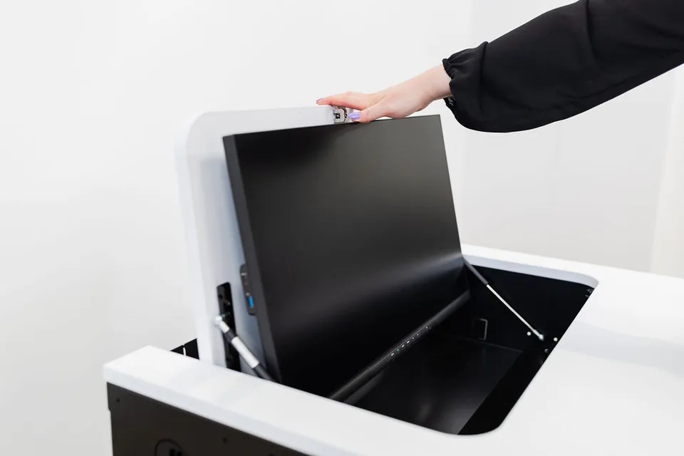 SMESK Flip von schräg oben: der Monitor steht an der aufgeklappten Klappe, die schwarze Wanne in der Tischplatte ist offen, eine Hand liegt auf der Klappe