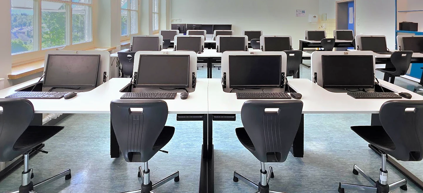 Derselbe Klassenraum als Computerraum: die Monitore sind aufgeklappt und stecken teilweise in der Platte, davor Tastatur und Maus
