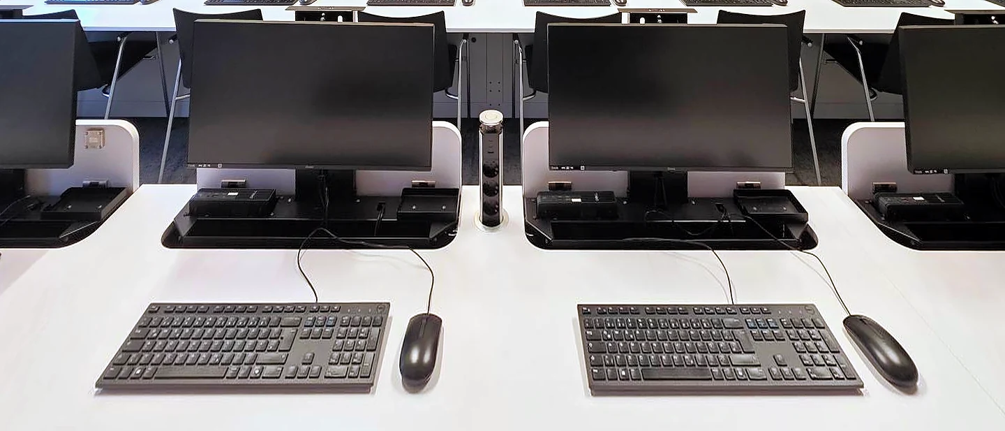Reihe von SMESK Lift Tischen von vorn: ausgefahrene Monitore, davor Tastatur und Maus, zwischen zwei Plätzen ein Steckdosenturm