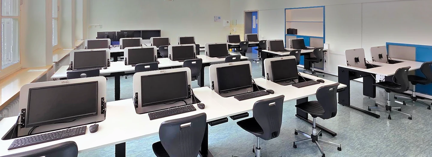 Klassenraum mit Reihen von SMESK Flip Tischen: die Monitore stehen aufgeklappt in den weißen Platten, davor Tastatur, Maus und Drehstühle