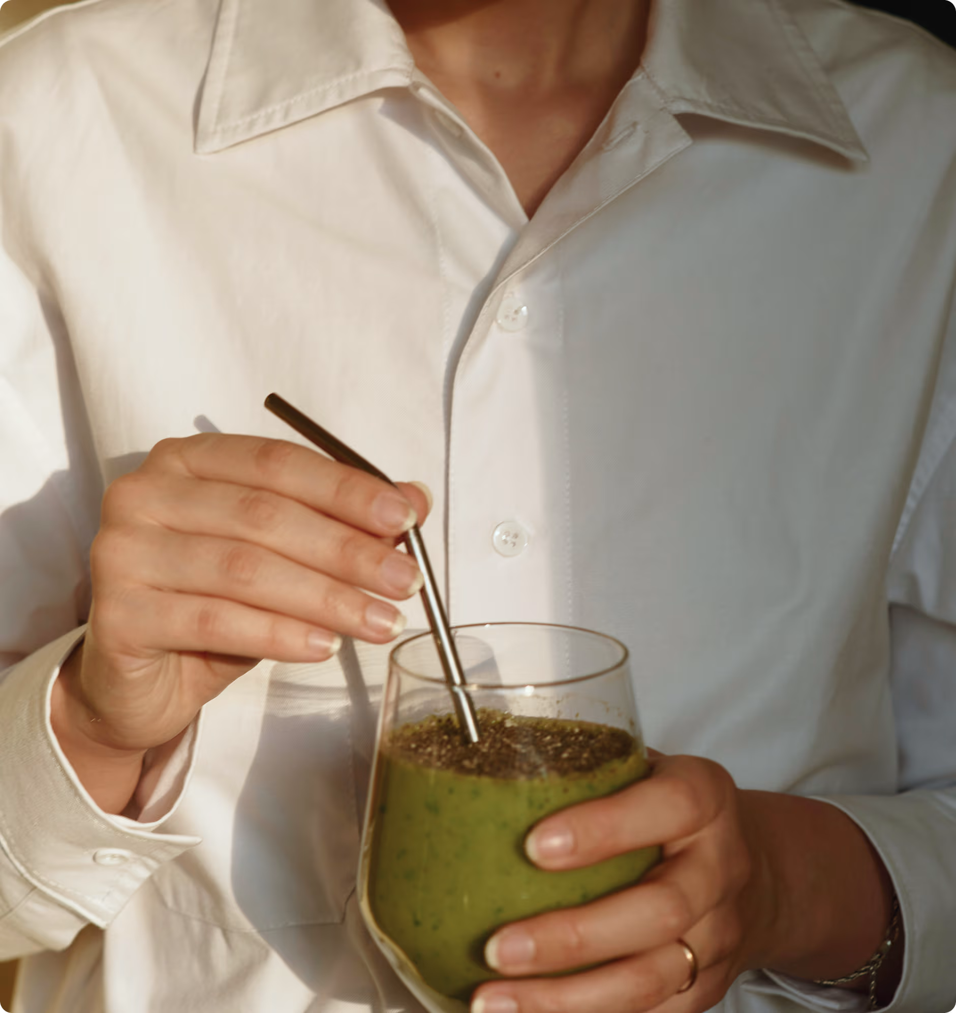 Une personne en chemise blanche tenant un verre de smoothie vert avec une paille métallique.