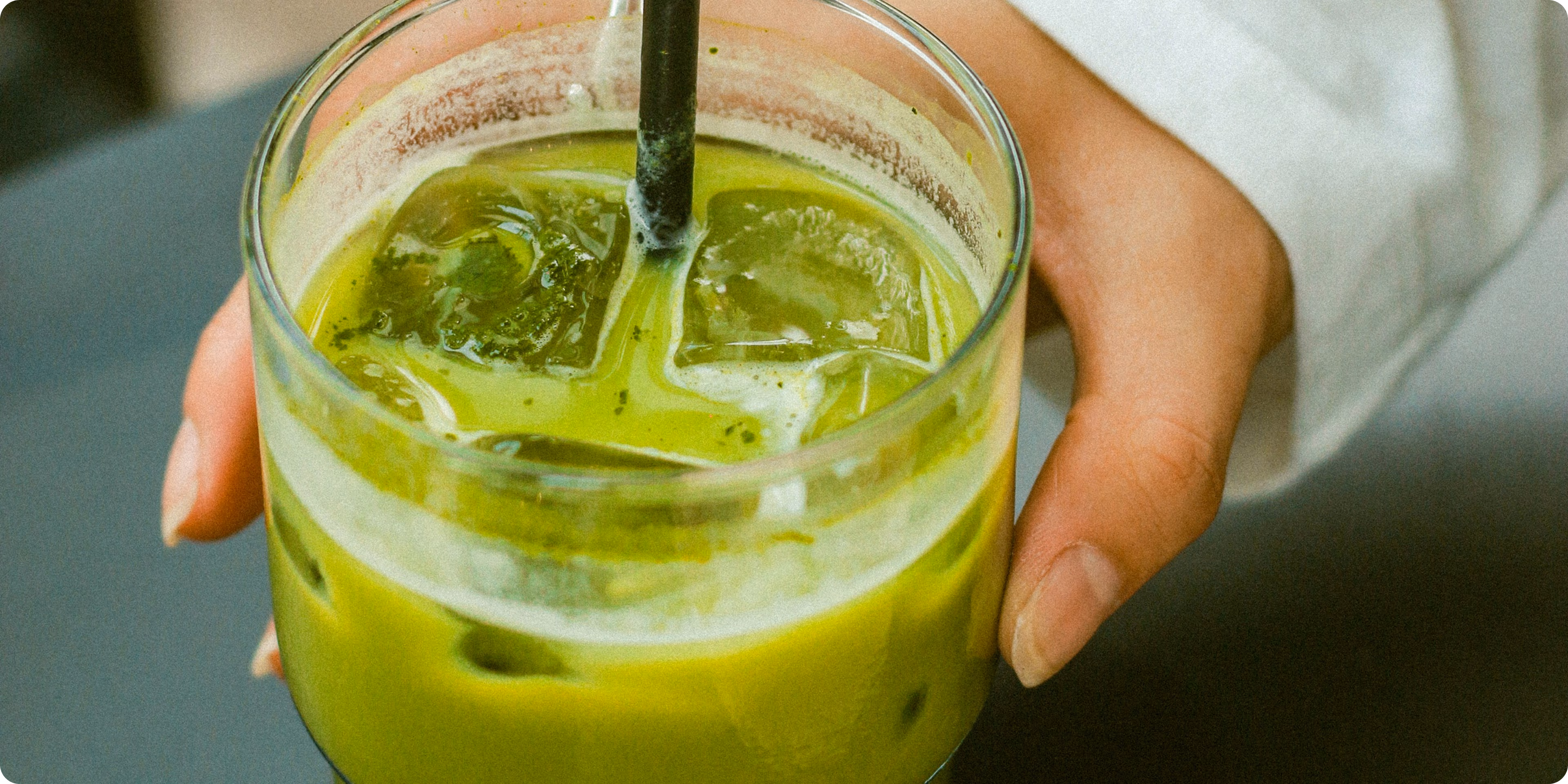 Main tenant un verre transparent rempli d'un thé vert glacé avec des glaçons et une paille noire.