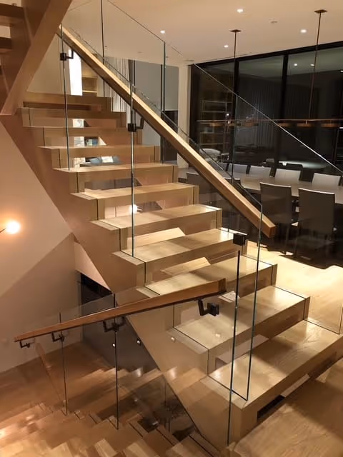 Modern wooden staircase with glass railings in a dining area with a large table and chairs visible through the glass.