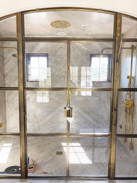 Spacious shower enclosure with large glass panels framed in gold, featuring a rain showerhead and a handheld shower wand.