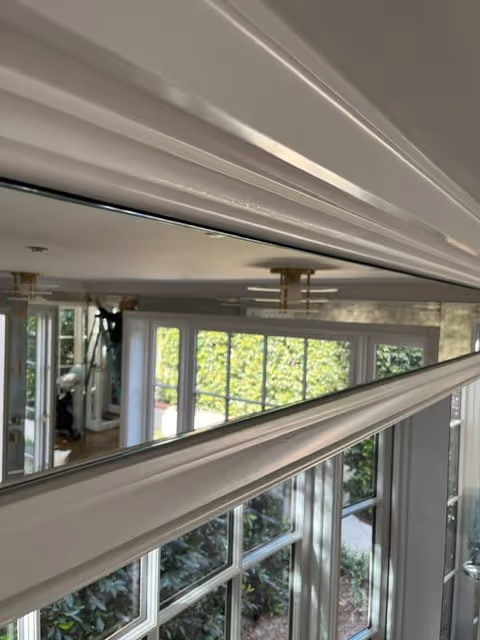 Close-up view of white crown molding inside a house with large windows and greenery outside.