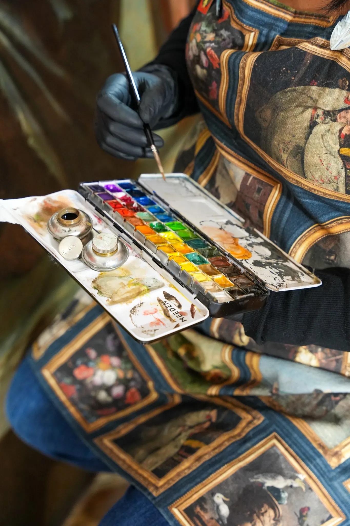 Person portant un tablier décoré tenant une palette de peinture aquarelle avec un pinceau dans une main gantée.