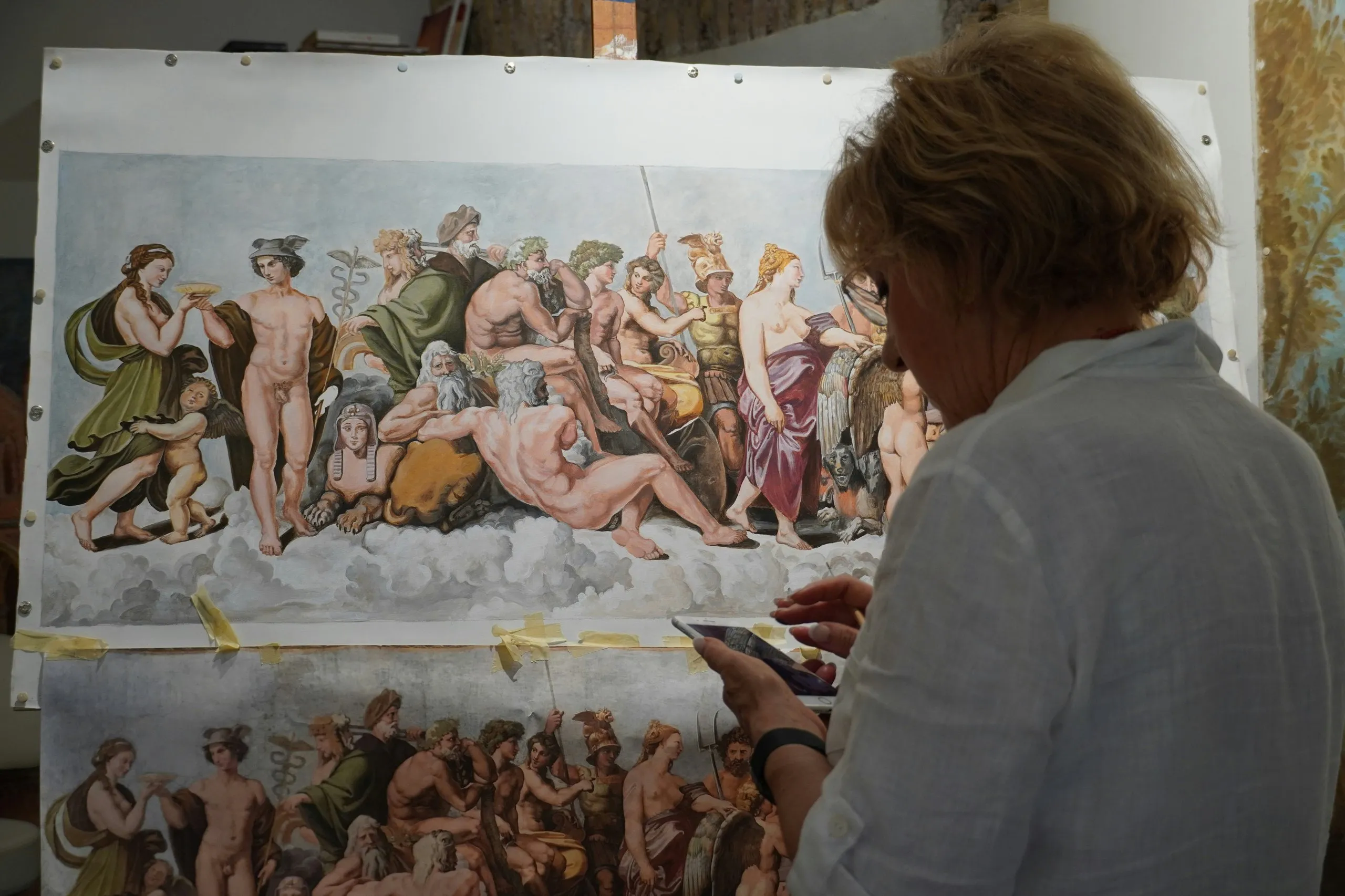 Personne tenant un téléphone devant une reproduction d'une peinture classique montrant plusieurs dieux et figures mythologiques sur un fond de nuages.