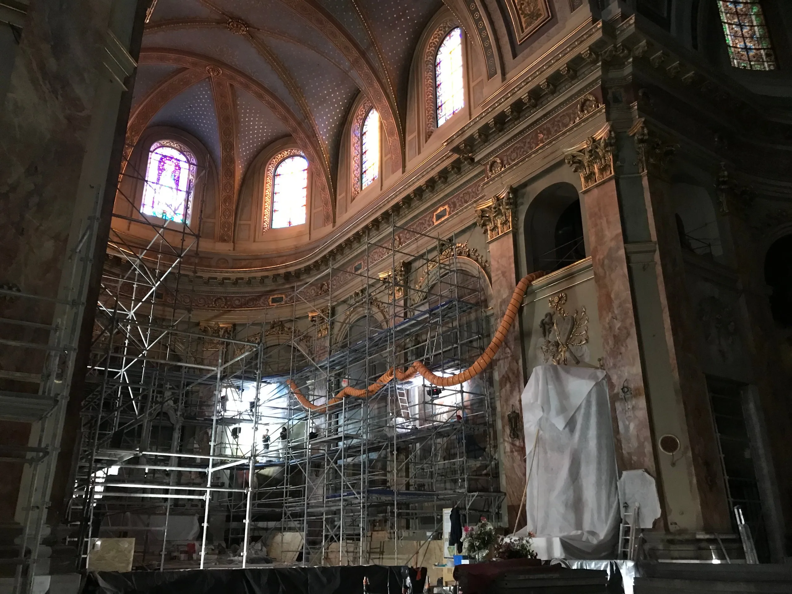 Intérieur d'une église historique avec des vitraux colorés, sous un plafond voûté orné, en cours de restauration avec des échafaudages métalliques et une statue couverte de tissu blanc.