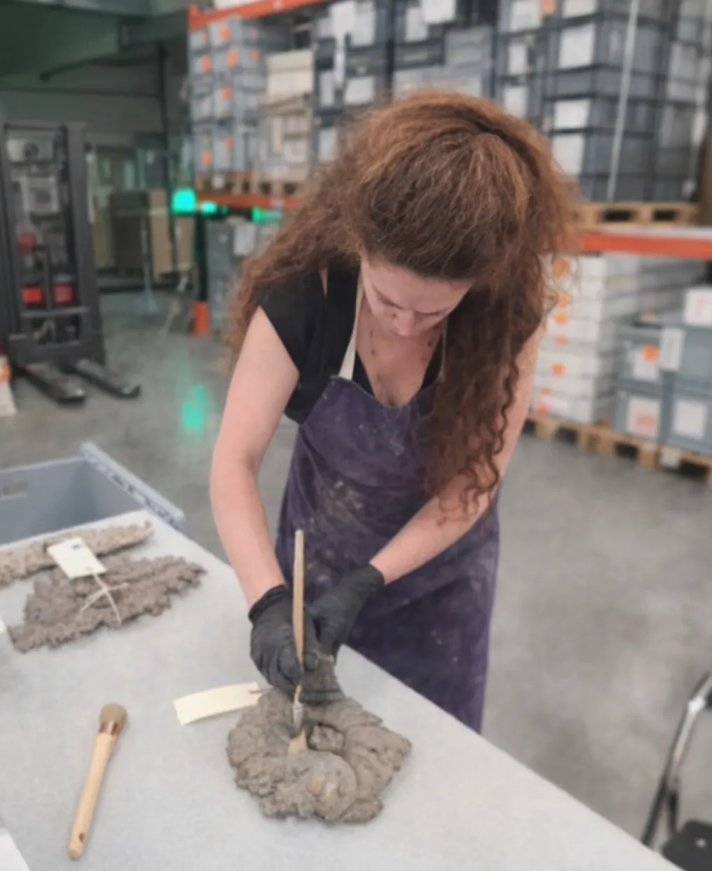 Femme avec un tablier violet sculptant un objet en argile sur une table dans un atelier.