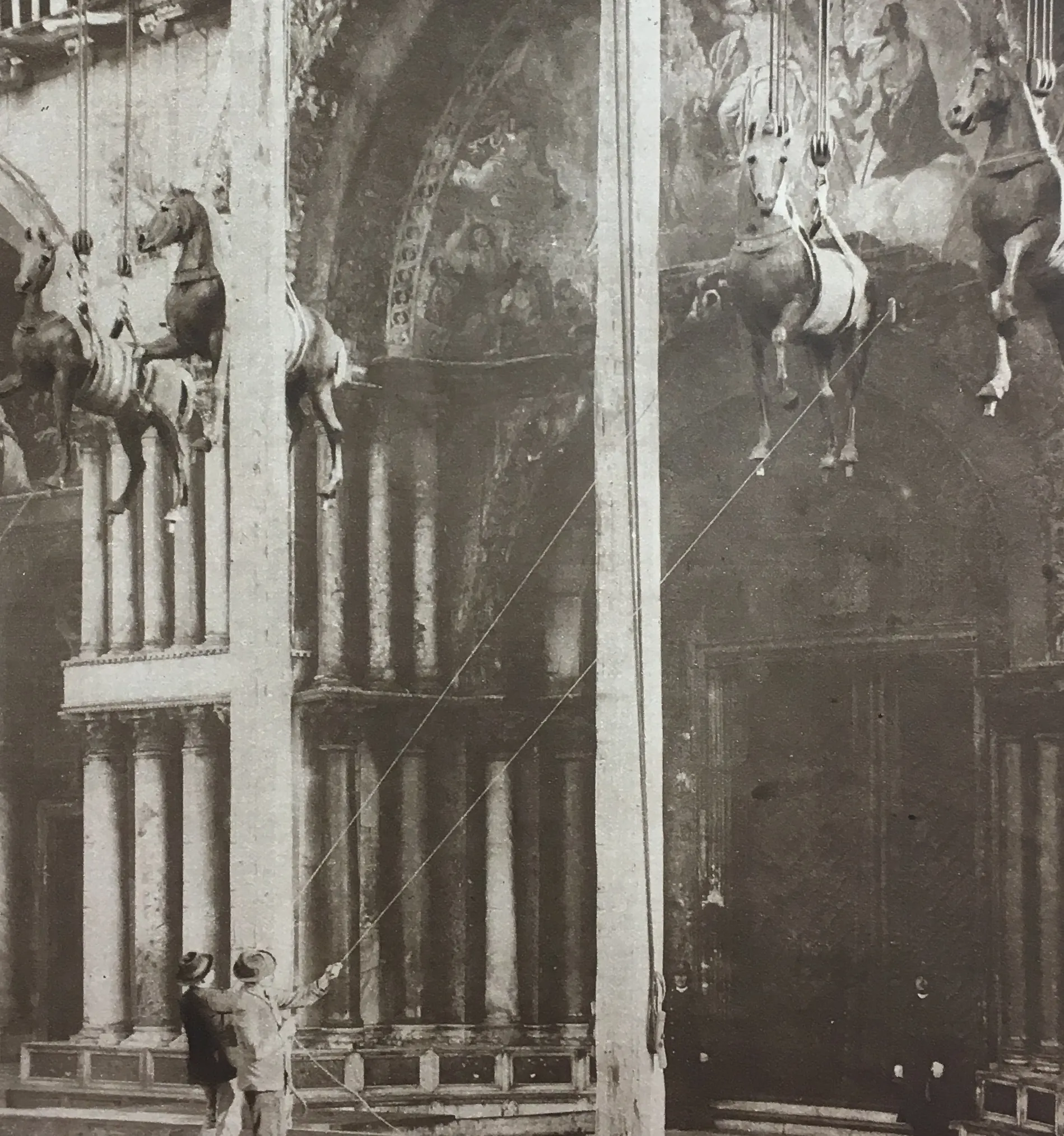Des personnes utilisent des cordes pour soulever plusieurs statues de chevaux suspendues dans un bâtiment historique avec des colonnes et des fresques murales.