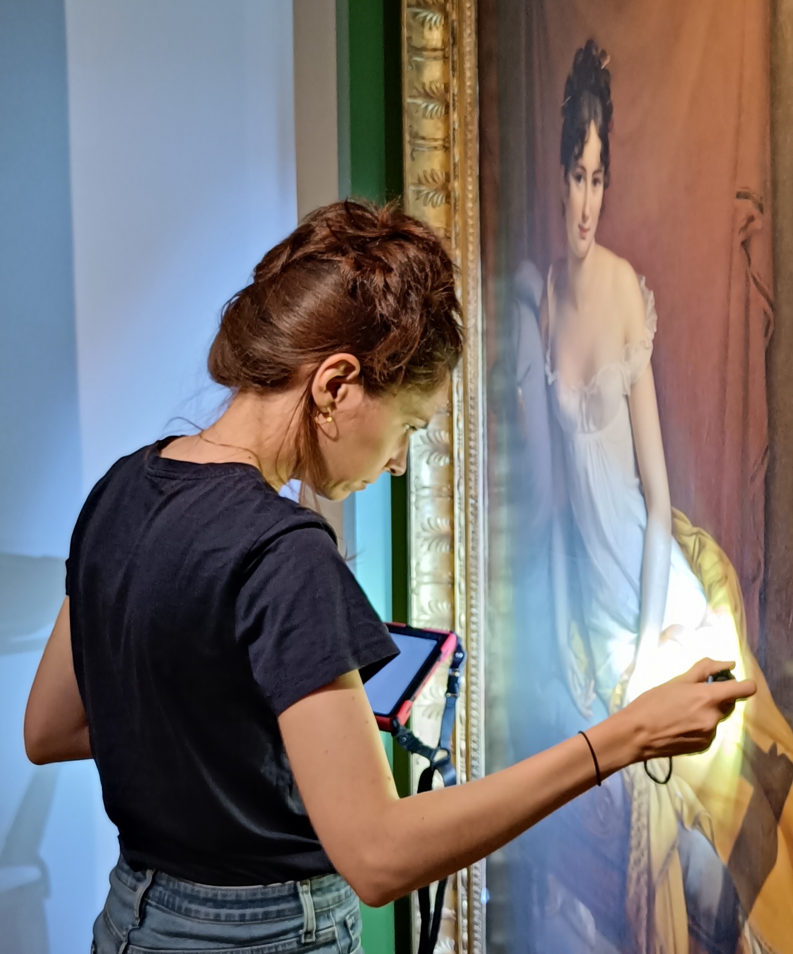 Une femme examine une peinture classique à la lumière d'une lampe de poche, avec une tablette suspendue à son poignet.