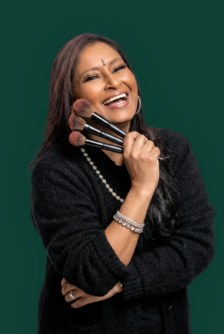 A joyful woman smiling warmly while holding three Kalay Beauty fluffy makeup brushes near her face. She is wearing a black sweater and silver jewelry against a clean green background.