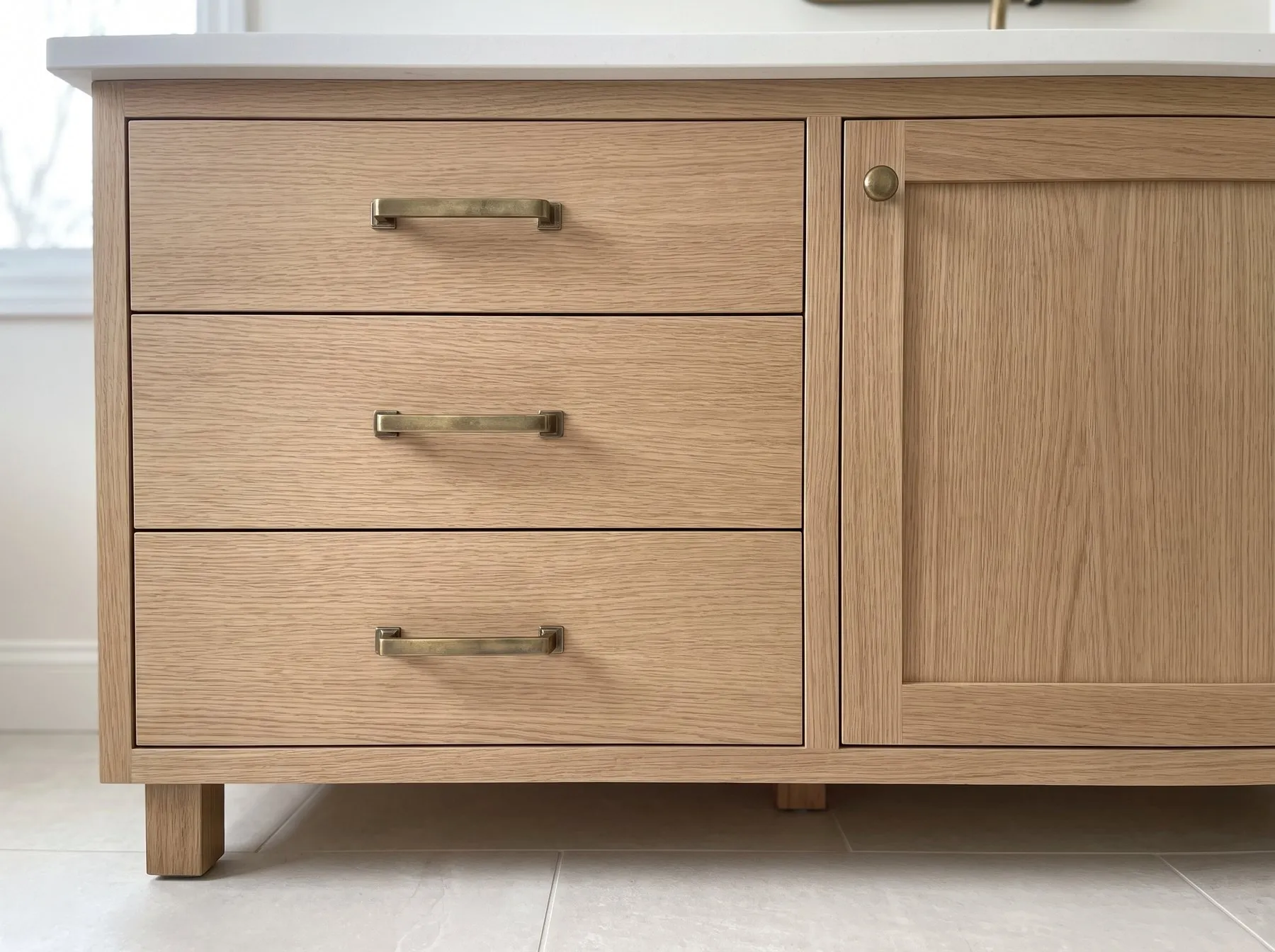 Close view of a natural oak vanity with three drawers, brass pulls and a white top, standing on short legs
