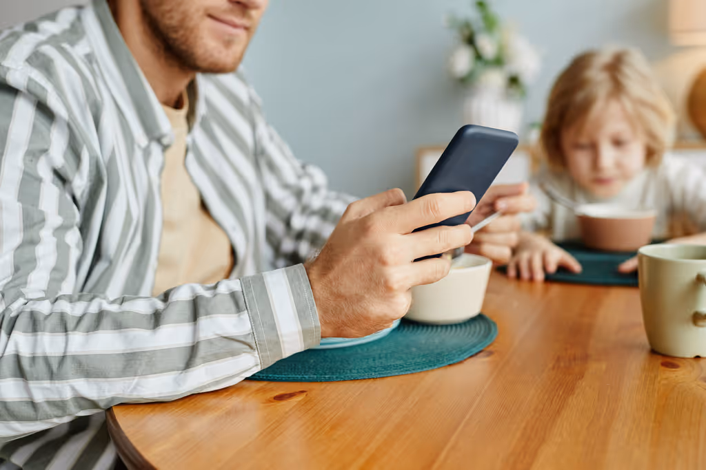 Man in striped shirt holding a smartphone at a dining table with a child eating cereal in the background.