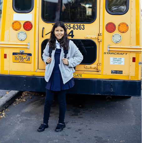 Young girl wearing a school uniform and backpack standing in front of a yellow school bus.