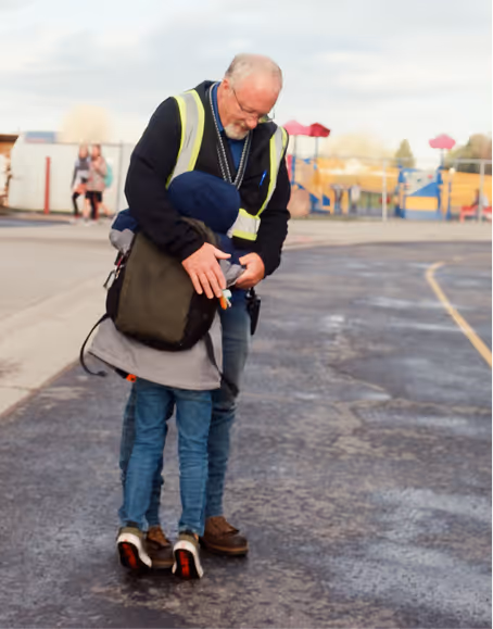 An elderly man in a reflective safety vest bending down to embrace a child wearing a blue hooded jacket and jeans on an outdoor paved area.