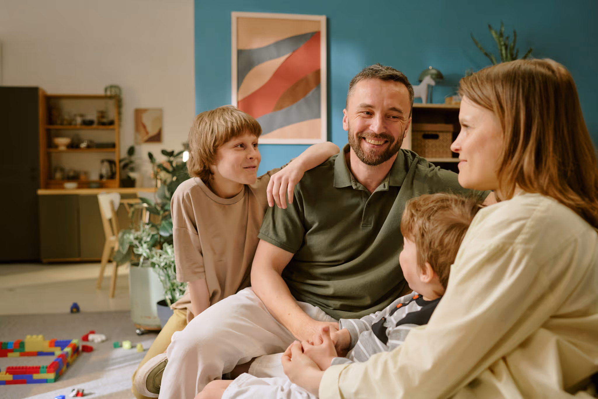 Smiling family of four sitting and talking together in a living room with colorful toys on the floor.