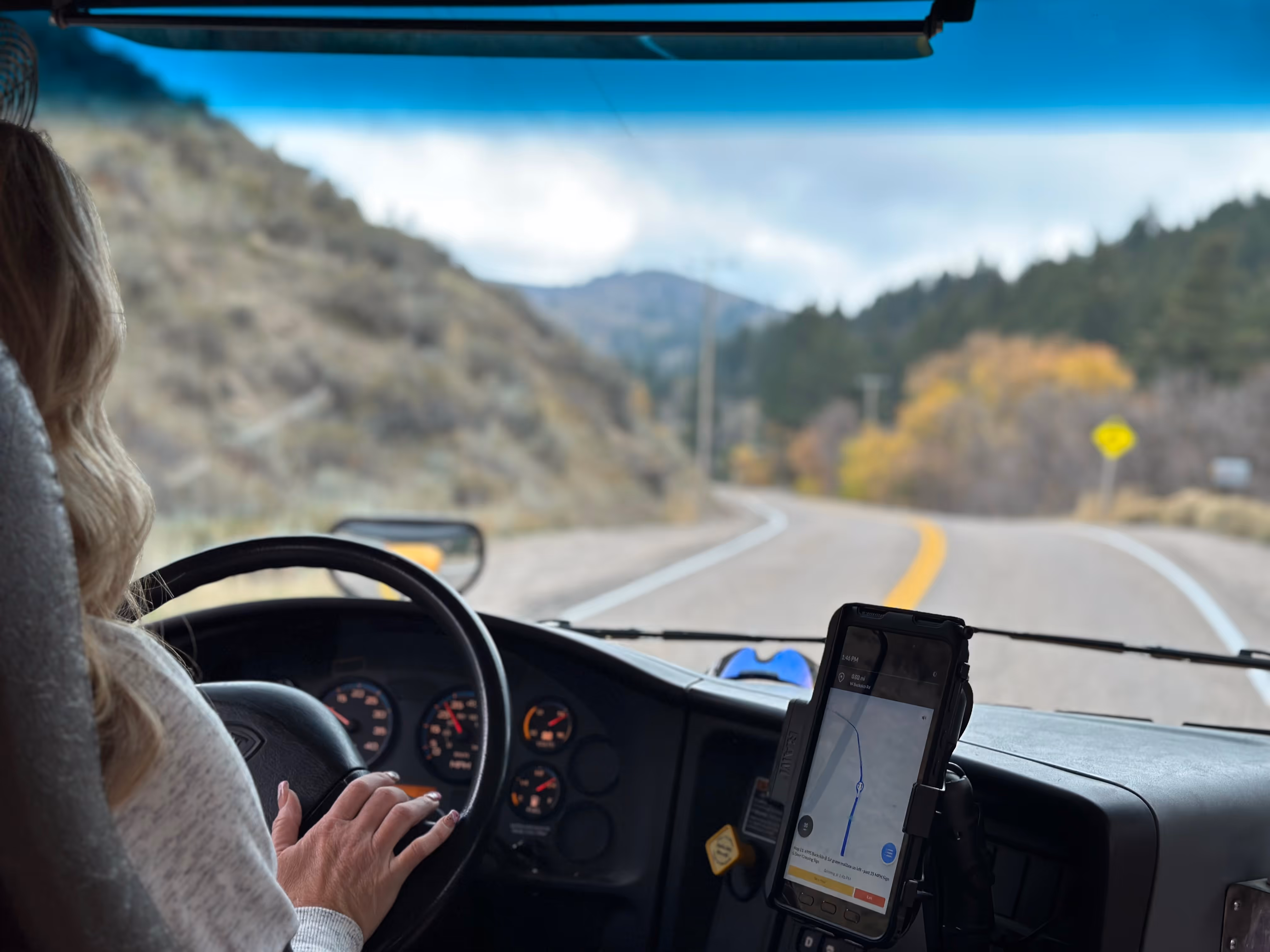 Person driving a vehicle on a winding road in a mountainous area with a GPS device mounted on the dashboard.