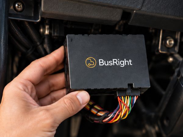 A hand holding the BusRight telematics device, a small black box with a wiring harness attached, in the footwell of a school bus below the dashboard.