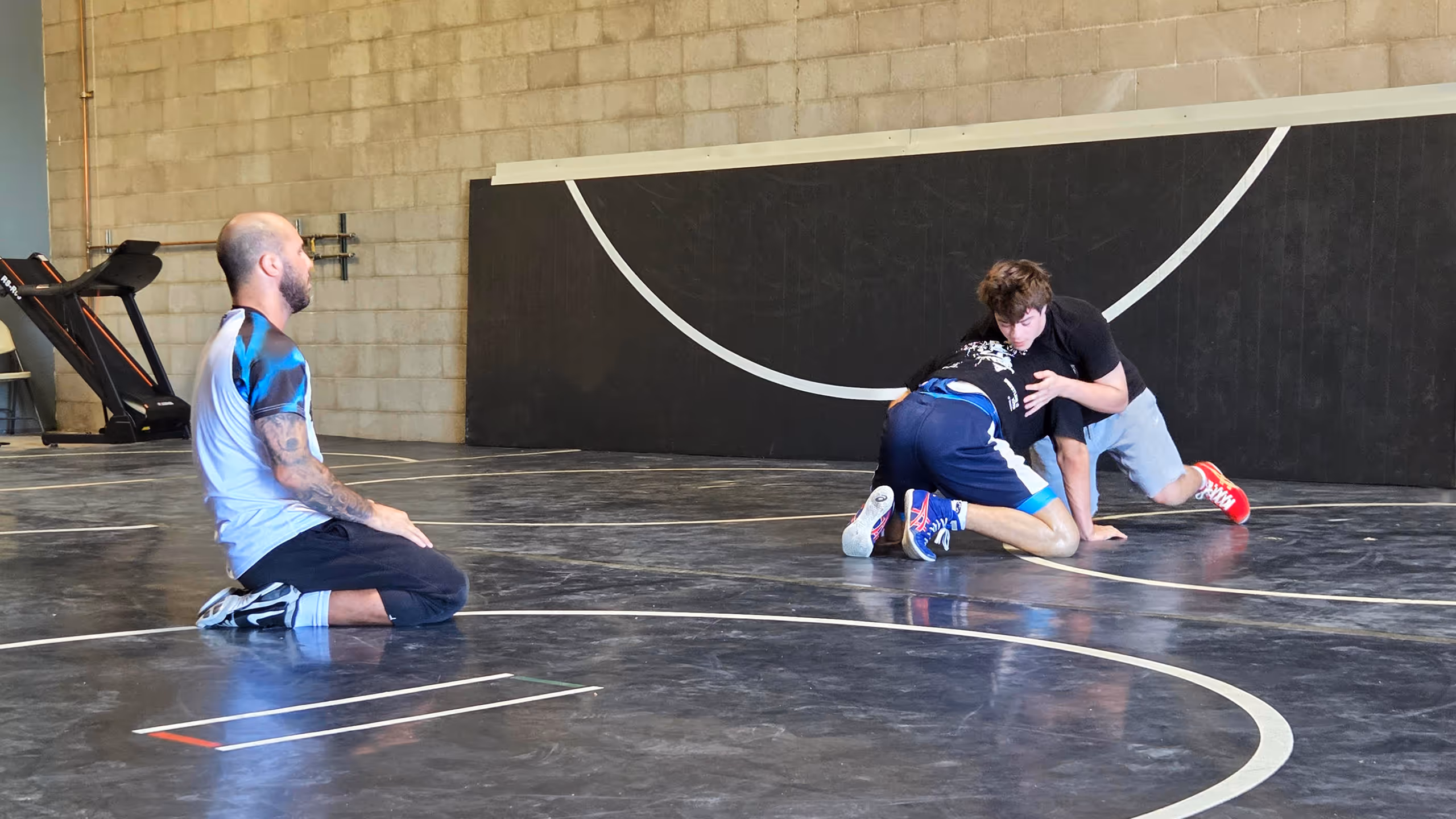 Two young men wrestling on a black mat in a gym while a coach with tattoos kneels nearby observing.