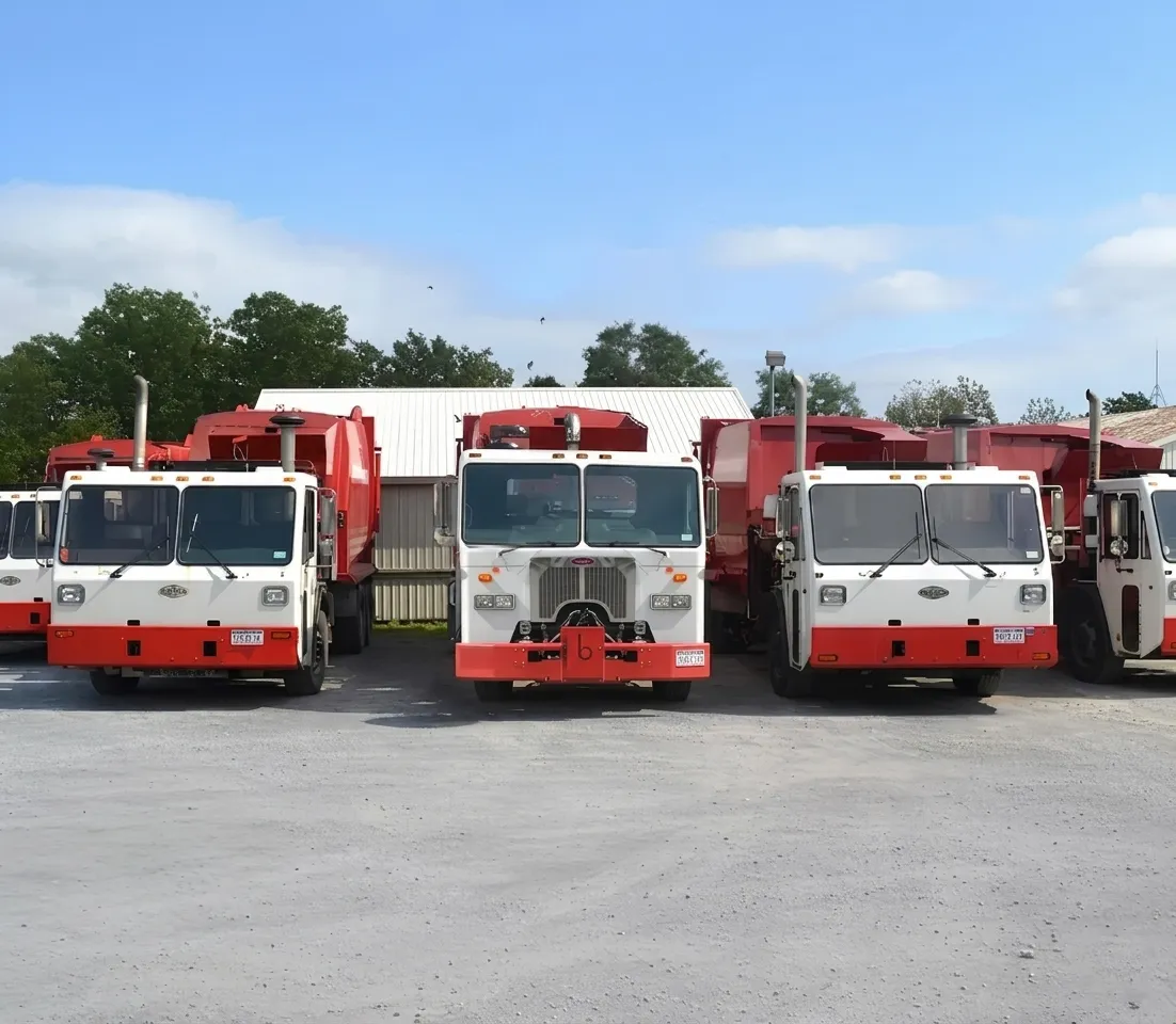 Fleet of red and white garbage trucks parked in a row on a gravel lot under a blue sky.