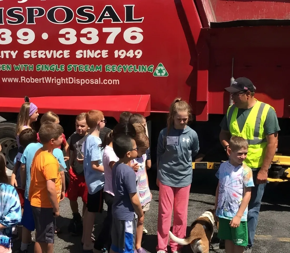 Group of children gathered outdoors near a red waste disposal truck, with a worker in a yellow safety vest and a dog.