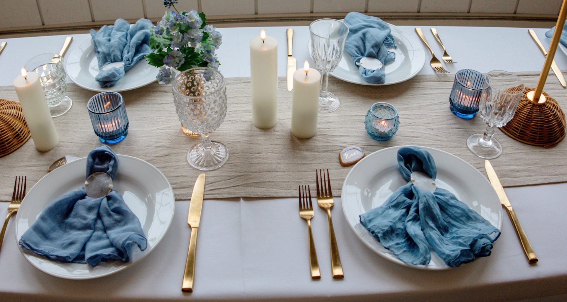 An elegant, coastal-themed tablescape with blue linens, gold flatware, and lit pillar candles for a curated event at Heritage Social in Douglas, Michigan.