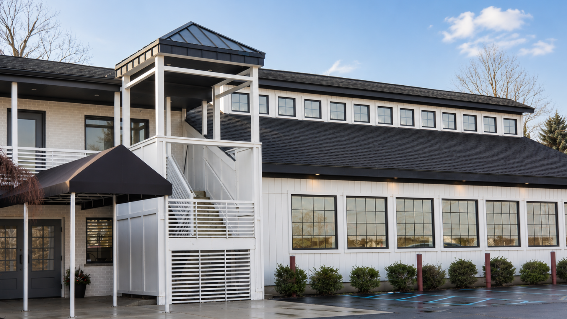 The exterior building facade of Heritage Social, an intimate event and wedding venue in Douglas, Michigan, featuring a modern white board-and-batten exterior, large black-trimmed windows, a dark shingle roof, and a black awning over the main entryway.