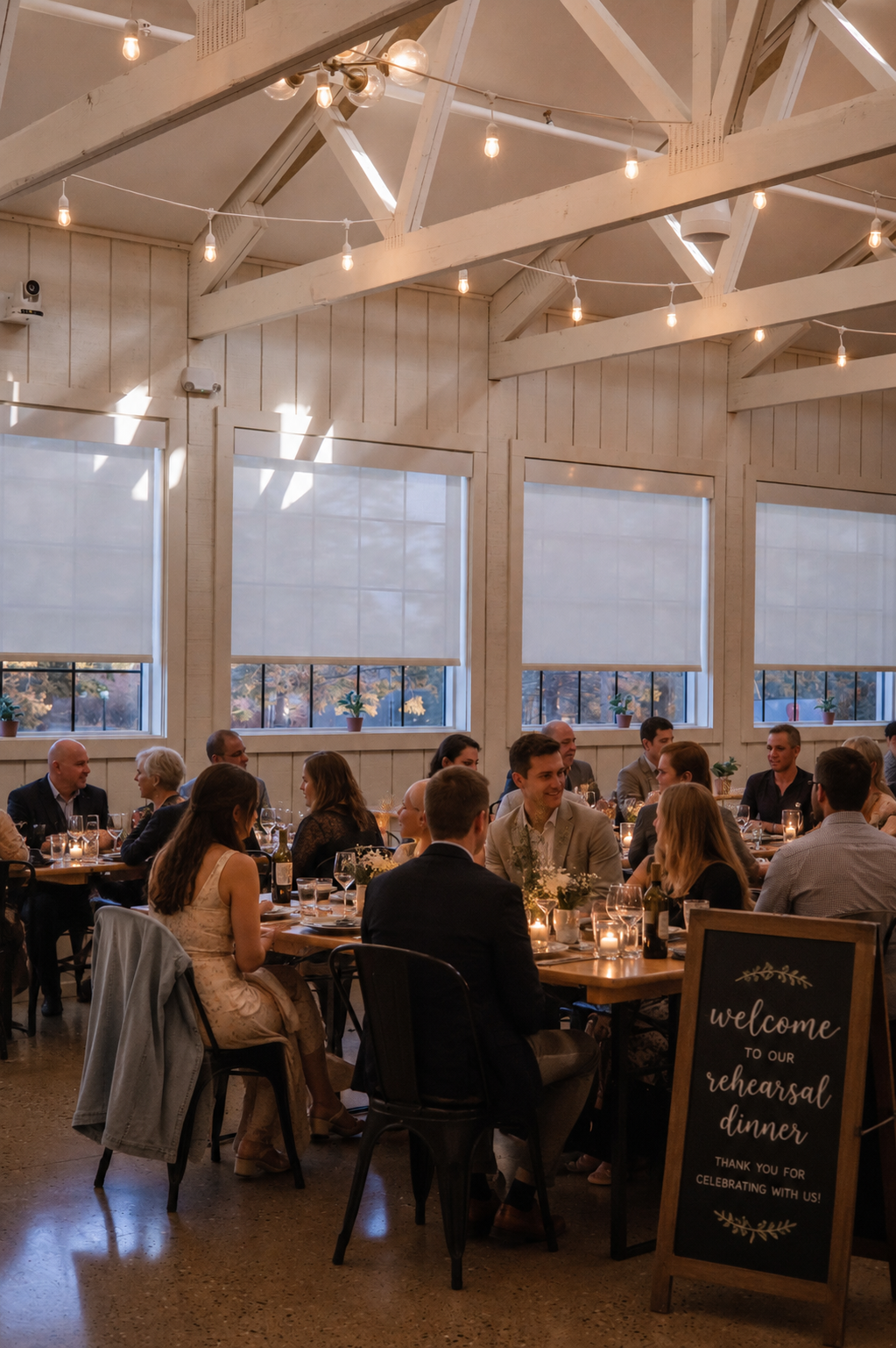 An active wedding rehearsal dinner celebration at Heritage Social in Douglas, Michigan, with guests seated at wooden tables under string lights and a chalkboard sign reading "welcome to our rehearsal dinner" in the foreground.