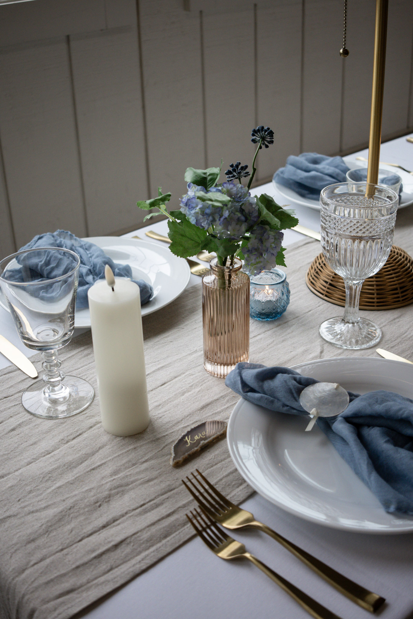 A beautifully styled coastal wedding table setting at Heritage Social in Douglas, Michigan, featuring a neutral linen runner, gold flatware, dusty blue cloth napkins, crystal glassware, white pillar candles, and a small vase of blue hydrangea flowers.