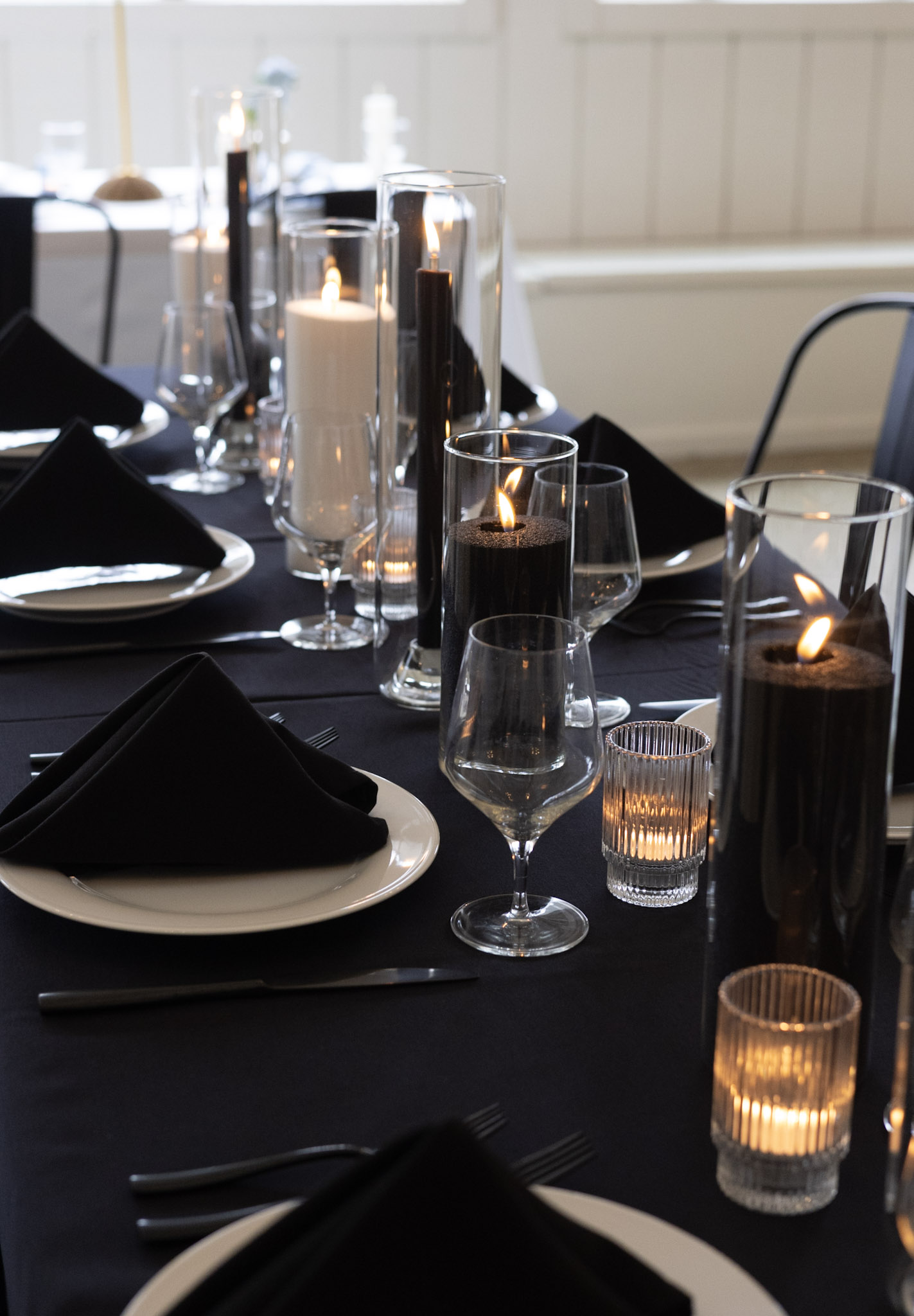 An elegant, moody tablescape designed for a luxury wedding rehearsal dinner or private celebration at Heritage Social in Douglas, Michigan, featuring black linens, black and white candles lit in glass cylinders, and white plates with folded black napkins.