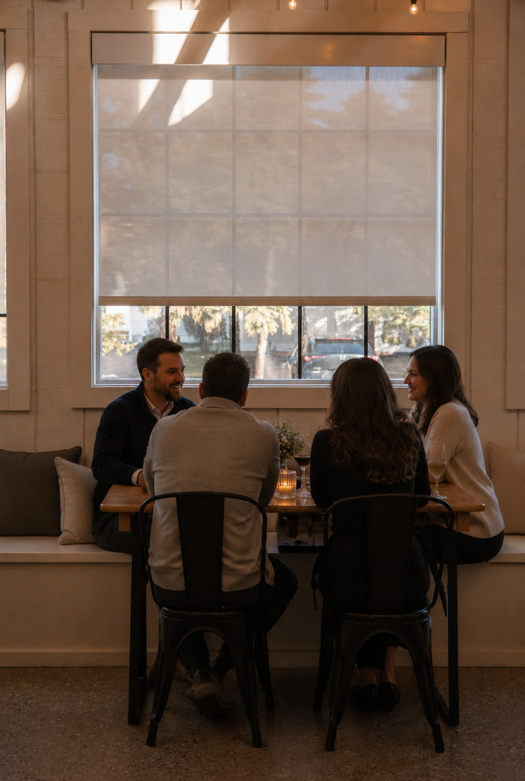 Four guests sitting at a wooden table next to a window with a sunshade pull-down blind, enjoying wine and conversation during a small private gathering at Heritage Social in Douglas, Michigan.