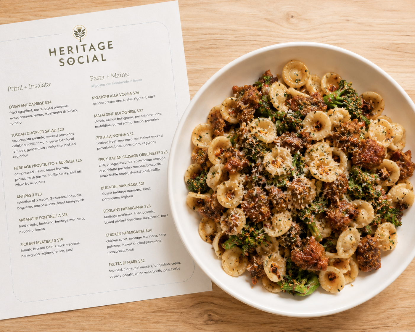 A printed Heritage Social event menu displaying "Primi + Insalata" and "Pasta + Mains" selections, sitting on a wooden table next to a large bowl of fresh orecchiette pasta with sausage and broccolini at an intimate venue in Douglas, Michigan.
