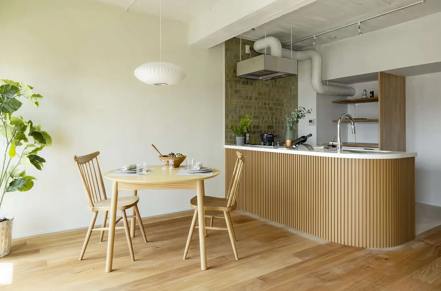 Modern kitchen and dining area with light wood table, two chairs, potted plant, and kitchen island with curved wooden paneling and stainless steel faucet.