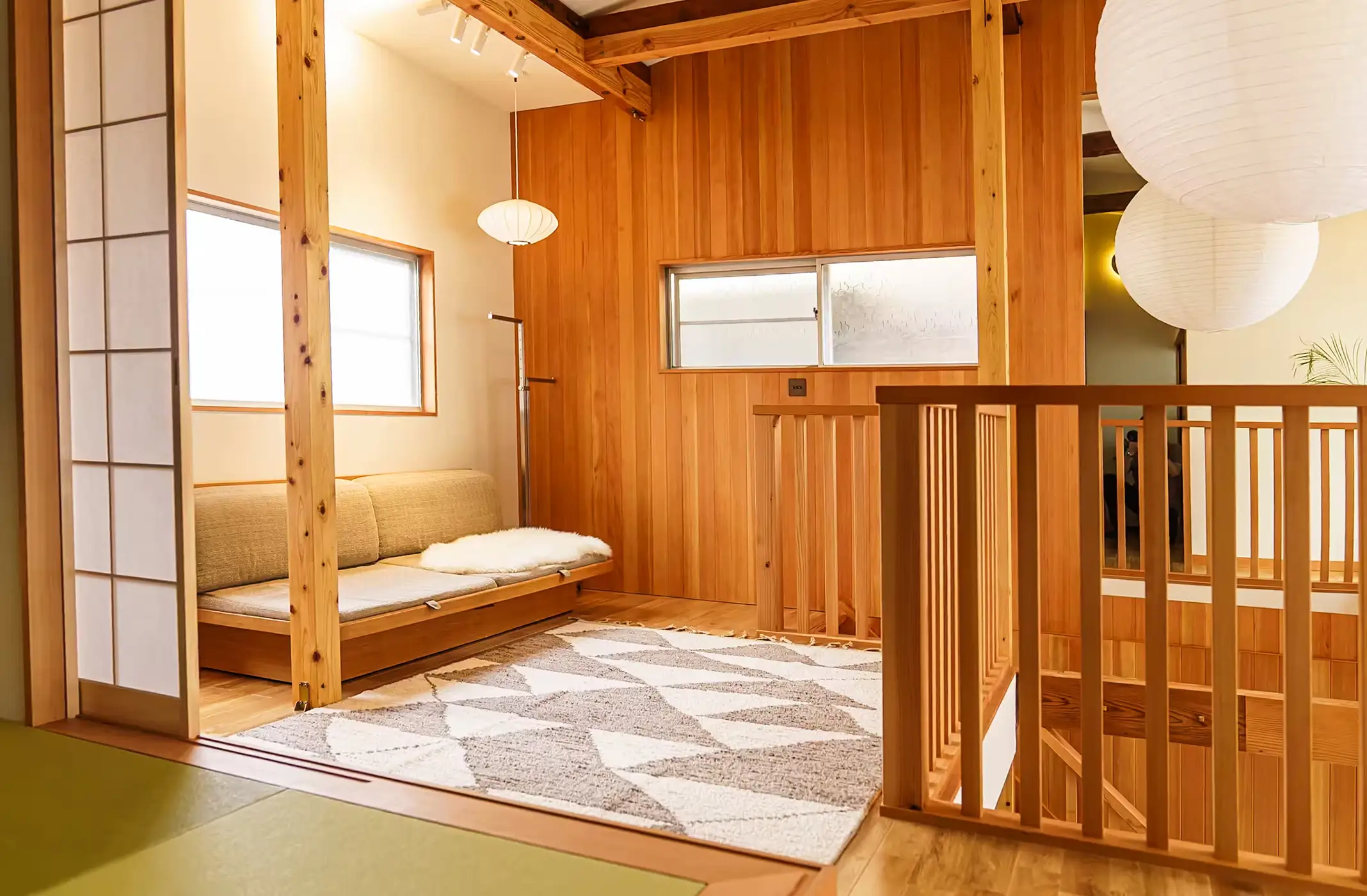 Cozy wooden loft space with beige sofa, geometric rug, paper lanterns, and wooden railings.