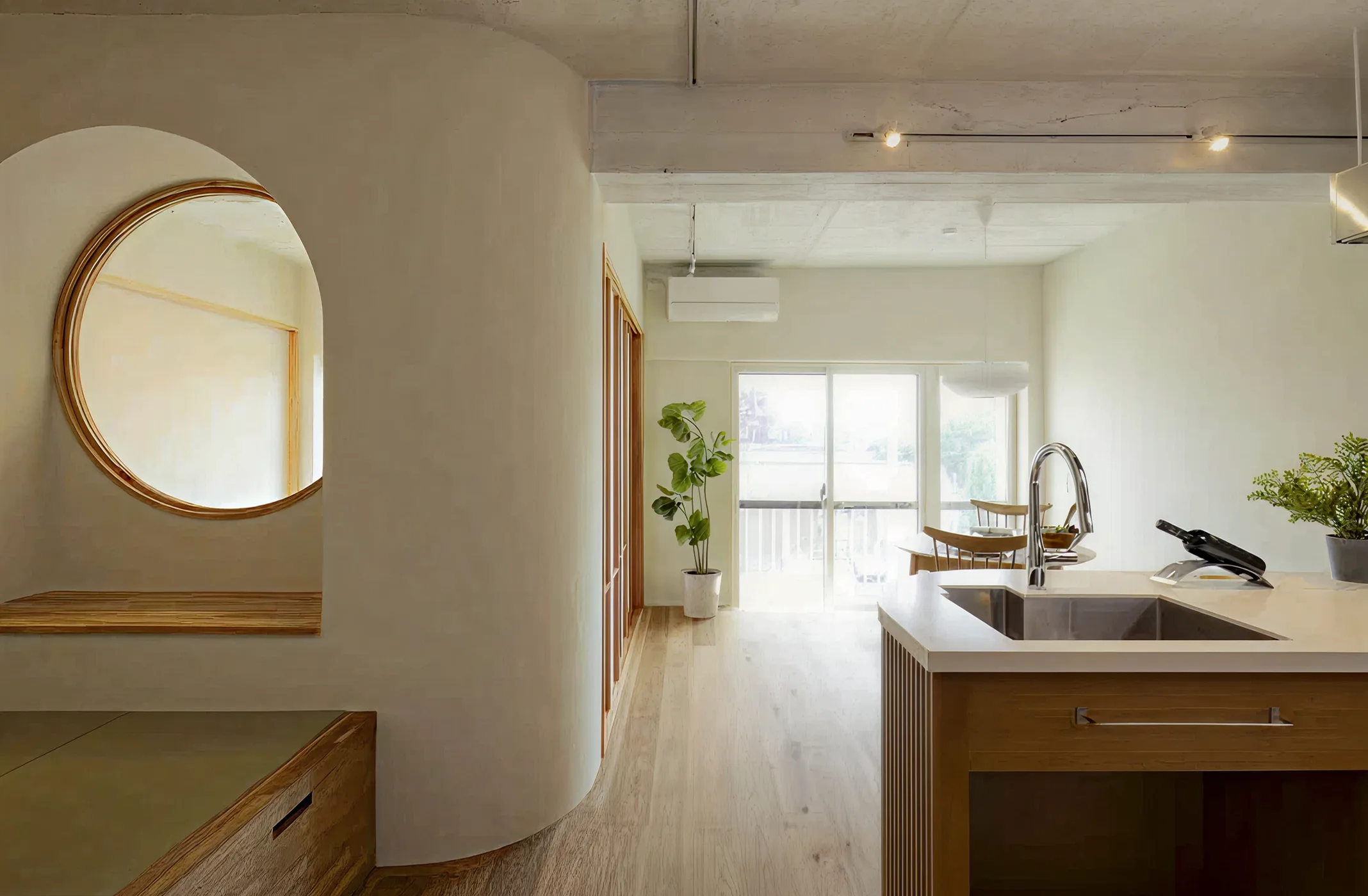 Minimalist kitchen and dining area with wooden floor, island sink, round mirror, and potted plants.