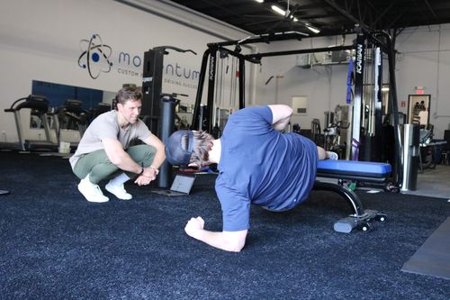 A man in a blue shirt and cap performs a side plank exercise on a gym bench while a trainer crouches nearby watching and encouraging him.