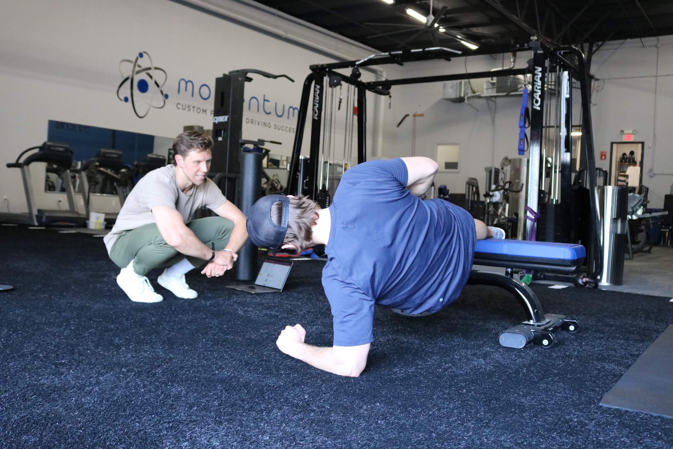 A man in a blue shirt and cap performs a side plank exercise on a gym bench while a trainer crouches nearby watching and encouraging him.