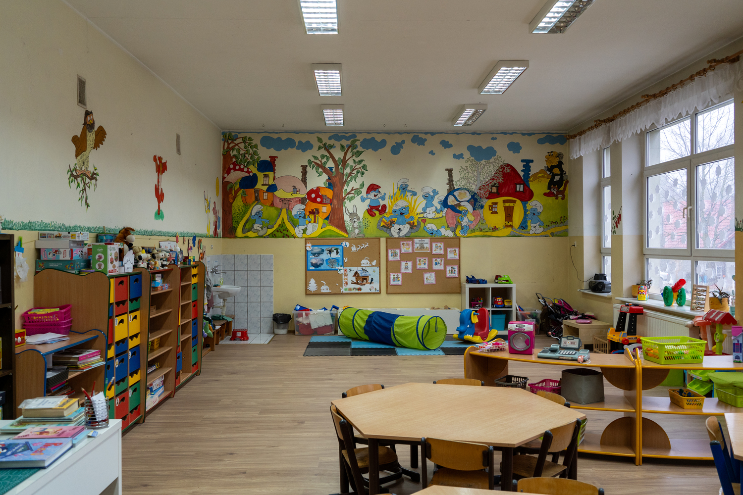 Young child drawing with markers at a classroom table with other children in the background.
