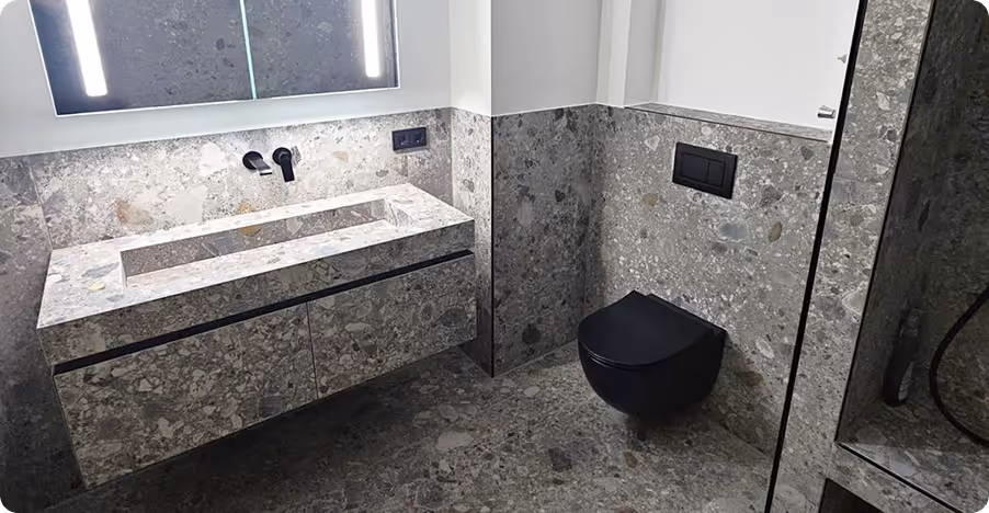 Modernes Badezimmer mit grauen Steinfliesen, wandhängendem schwarzen WC und Waschbecken mit beleuchtetem Spiegel.