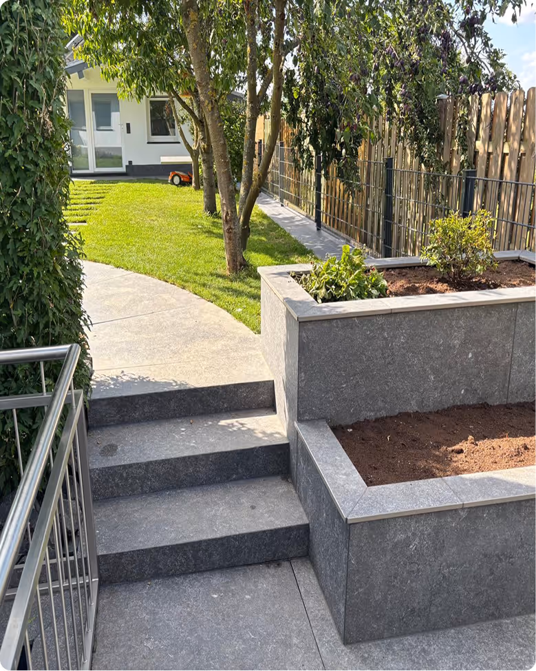 Moderner Garten mit grauen Steinstufen, einem Hochbeet mit Erde und Pflanzen, grünem Rasenteppich und Bäumen vor einem weißen Haus.