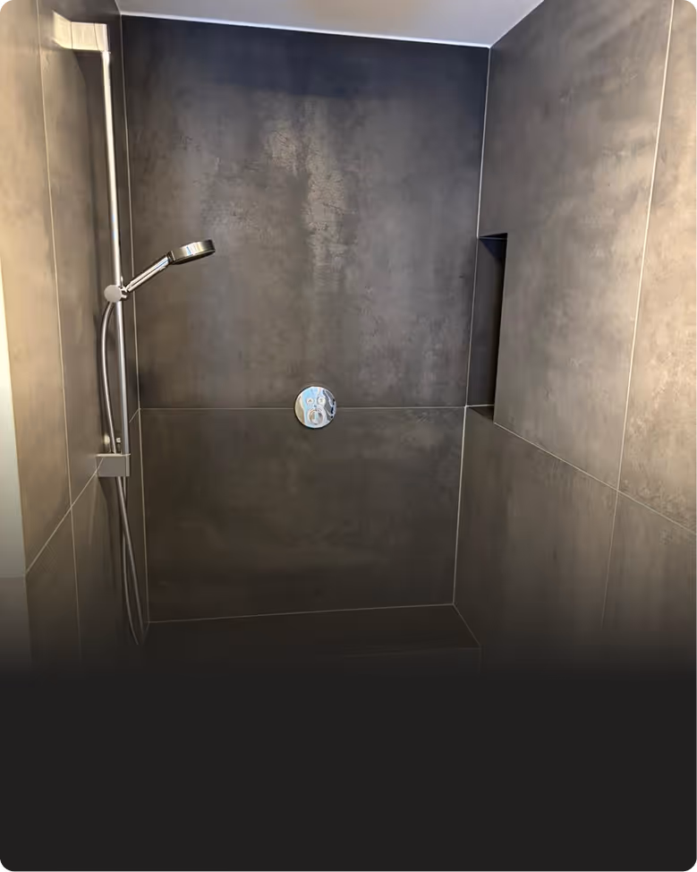 Moderne Duschkabine mit dunkelgrauen Wandfliesen, verstellbarem Duschkopf und eingebauter Ablage.