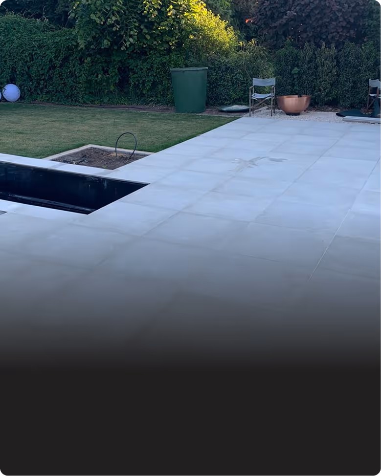 Garten mit einem rechteckigen Pool ohne Wasser, einem gepflasterten Bereich, zwei Gartenstühlen, einem grünen Abfalleimer und dichter Bepflanzung im Hintergrund.