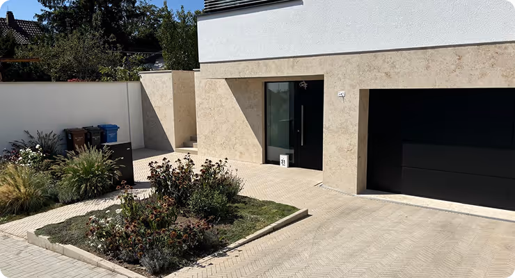 Vorgarten mit Blumenbeet, Pflastersteinen und moderner Hausfassade mit schwarzer Haustür und Garage.