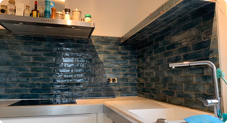 Moderne Küche mit blauen glänzenden Wandfliesen, Kochfeld, Dunstabzugshaube, Spüle und verschiedenen Gewürzflaschen oben.