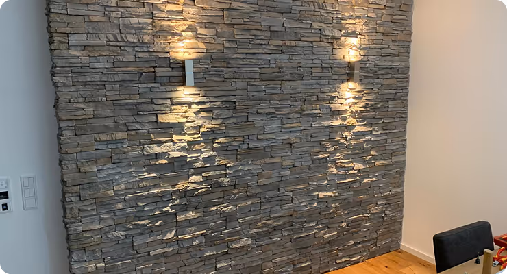 Innere Steinwand mit zwei modernen Wandlampen, die warmes Licht nach oben und unten strahlen, und Holzboden darunter.