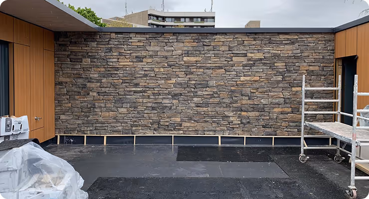 Rohbau eines Außenbereichs mit einer Wand aus natursteinähnlichen Ziegeln, Holzwänden links und rechts sowie Baugerüsten.