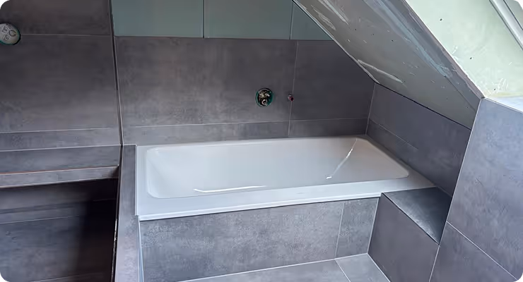 Modern bathroom with a white bathtub installed under a slanted ceiling, surrounded by gray tiled walls and floor.