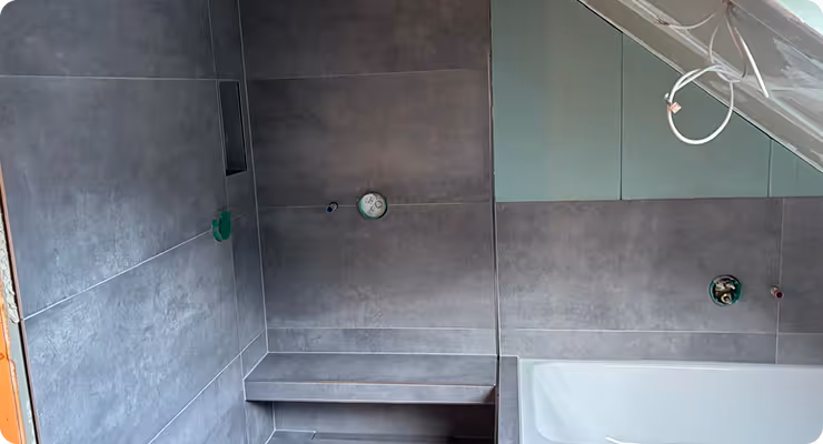 Graue geflieste Badewanne mit eingebauter Bank, unvollendete Installation mit sichtbaren Rohranschlüssen und weißem Deckenlichtkabel.